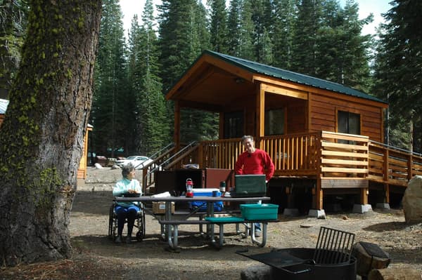 A woman sits in a wheelchair and a man stands at a picnic table backed by a small, wooden cabin. - NPS Photo
