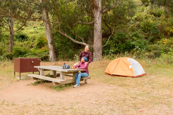 Campers sit at a picnic table next to a food storage box with their tent behind them. - NPS Photo